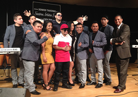 The hosts and artists of El Barrio también Canta pose for a picture at the end of the show on stage, many of them are smiling.