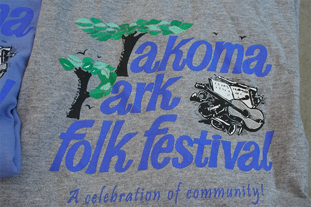 A gray shirt with a design for Takoma Park Folk Festival. The words are in blue and the T and P are trees. There is also a violin and other instruments to the left, and underneath are the words: A celebration of community!
