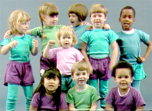 A group of 9 preschool children, with six standing, and three sitting. There are talking and looking at the camera, dressed in various shades of green, blue, and purple.