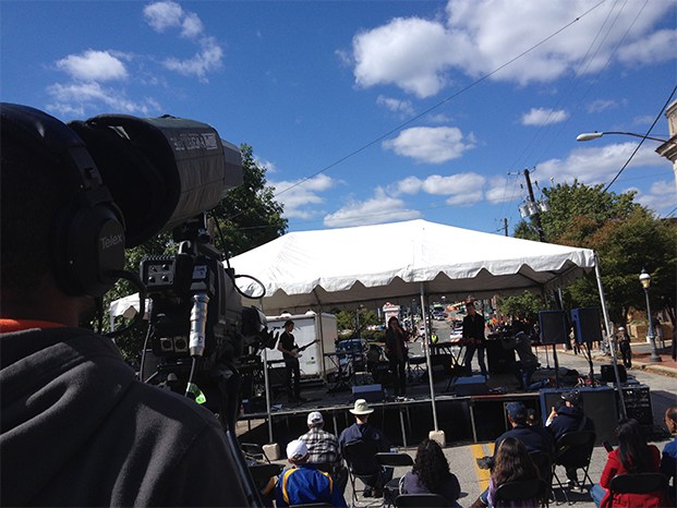 A camera operator is seen from behind filming a stage at the Street Festival on a bright day with a few clouds in the sky.