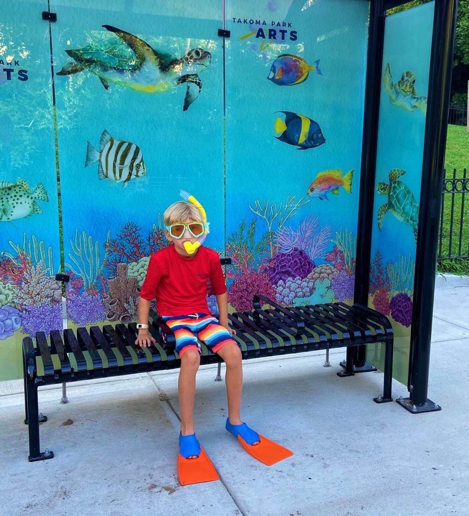 Boy in snorkel gear sits at newly installed bus stop with tropical fish decals. 